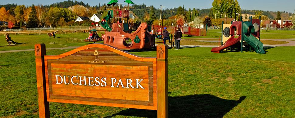 Duchess Park playground with sign that reads "Duchess Park" in foreground. Playground includes a pirate ship, climbing tower, and toddler playground.